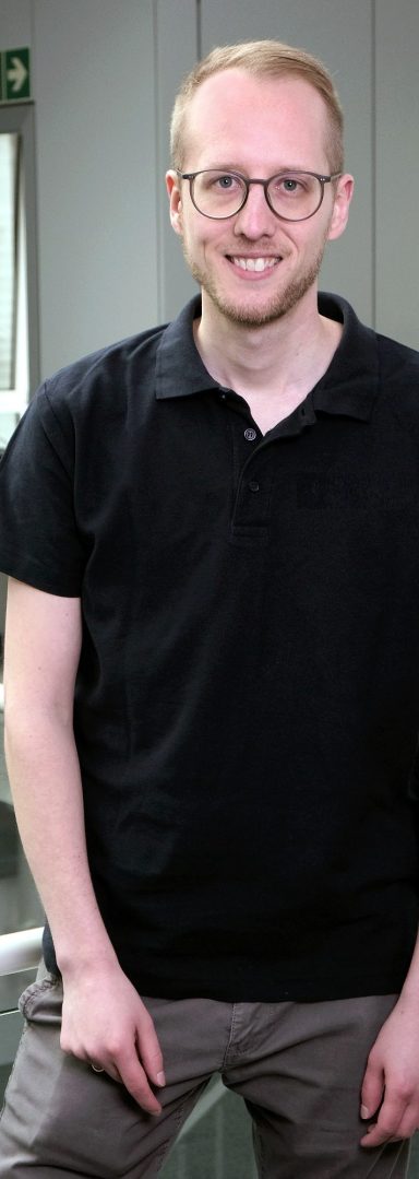 Lächelnder Mann mit Brille in einem hellblauen Hemd, steht in modernem Büro.