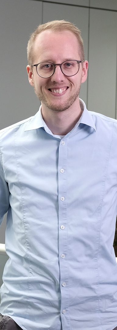 Lächelnder Mann mit Brille in einem hellblauen Hemd, steht in modernem Büro.